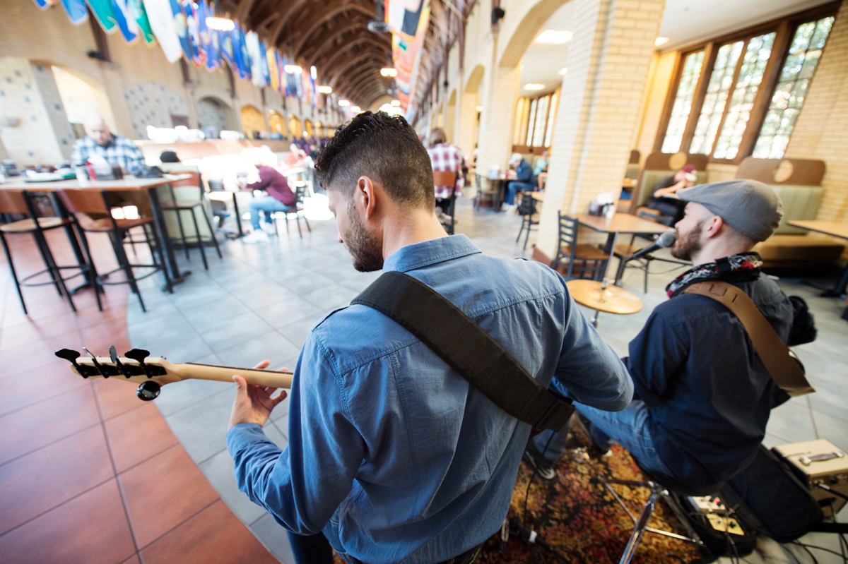 A musical duo plays to lunchtime diners at Perry Cafeteria, with their backs to the camera and Perry&amp;amp;#039;s flags in the background.