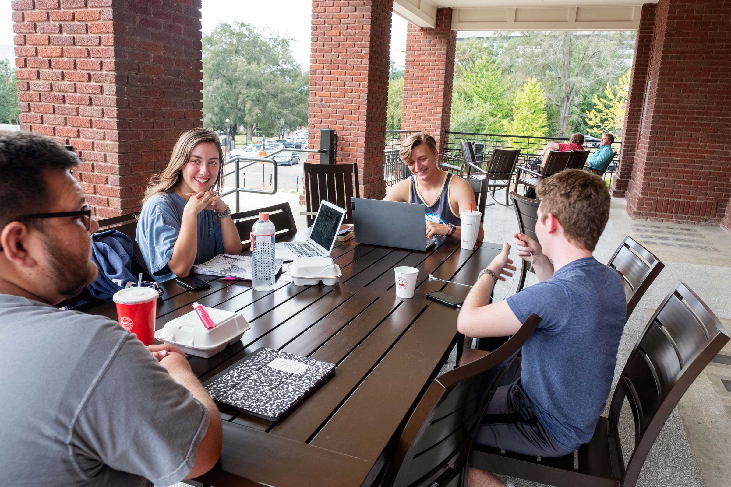Four students chat and eat lunch between classes at a table on the YMCA&amp;#039;s porch.