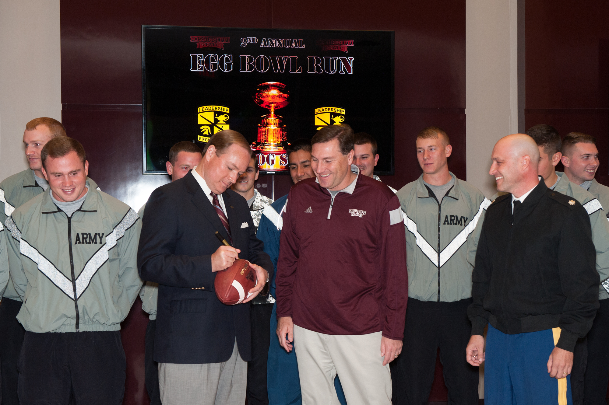 MSU President Mark E. Keenum, front left, head football coach Dan Mullen and Lt. Col. Brian Locke, the university's professor of military science and battalion commander, take turns signing the football that Army and Air Force ROTC cadets from MSU and Ole Miss will carry during Monday's second Egg Bowl Run.