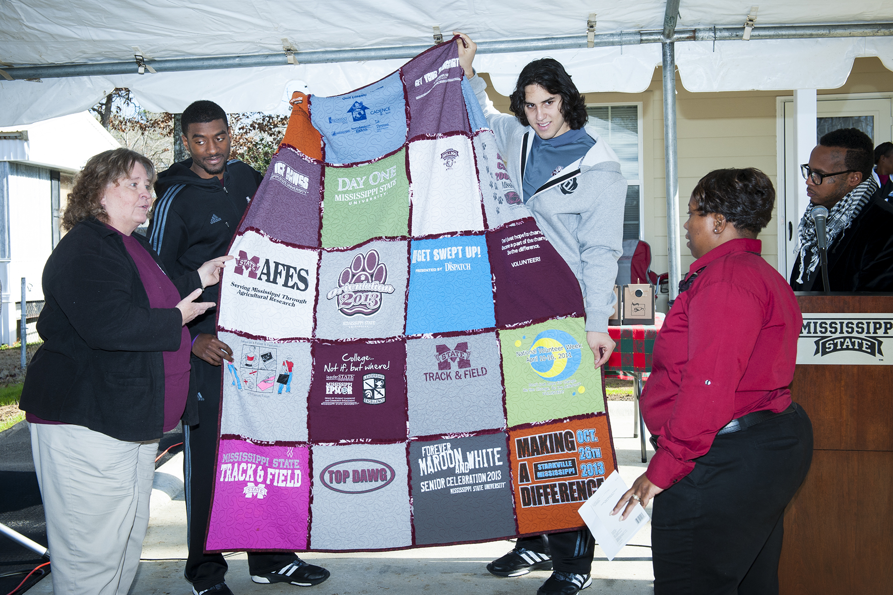 Starkville resident Dot Livingston, left, presents a quilt made from MSU T-shirts donated by Maroon Edition Habitat for Humanity volunteers to new homeowner Angela Lindsey, right. MSU student-athletes Jeffery Johnson and Johnny Zuppardo display the quilt during the home dedication ceremony Dec. 16.